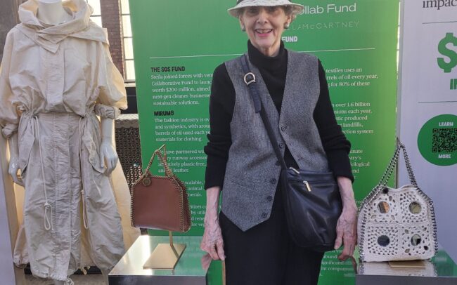 Laudato Si member at an exhibit booth on making fashion more environmentally sustainable. She is smiling and wearing a black and gray outfit and an ivory hat. The booth shows purses and a coat made to be more sustainable.