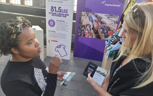 Climate Fair fashion waste exhibit booth. A Laudato Si member, a young Black woman with her hair tied back, talks with the booth presenter.