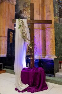 Photo of a large wooden cross in front of the columns at St. Francis. The cross is draped in white cloth and green vines. The base of the cross is draped in purple.